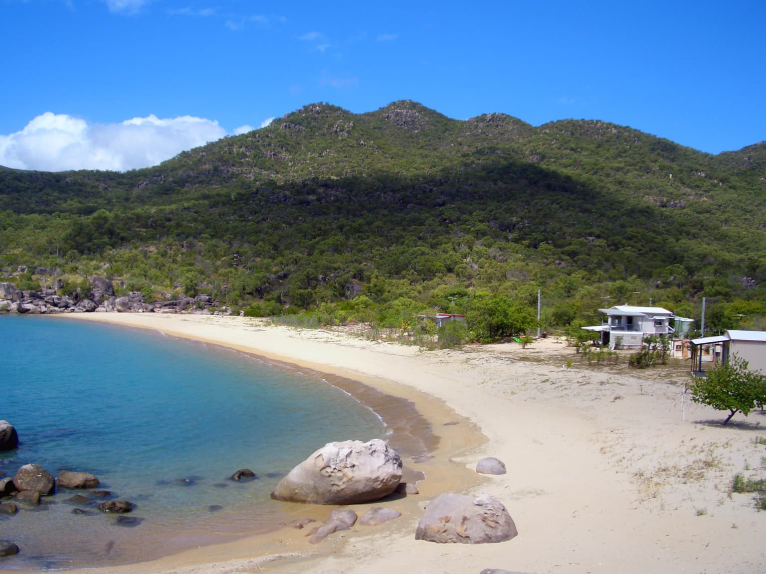 Playa arenosa con aguas turquesas, grandes rocas, terreno montañoso verde y algunos edificios bajo un cielo azul despejado