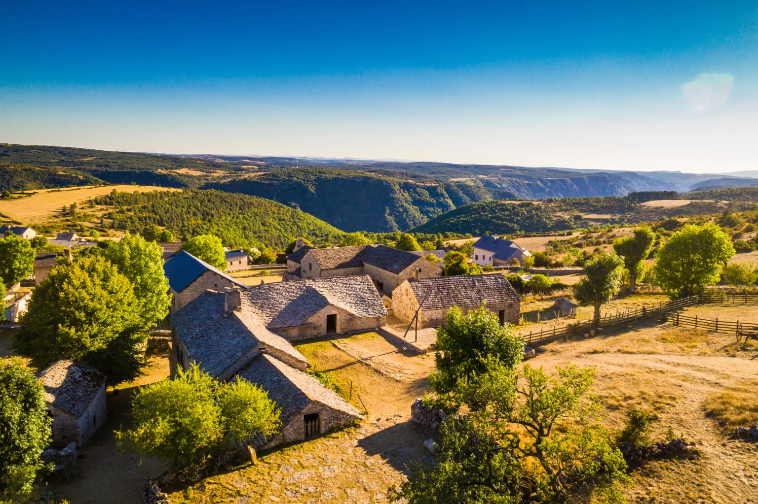 Stone farmhouse complex with stone roofs surrounded by trees and open fields, overlooking rolling hills and valleys under clear blue sky