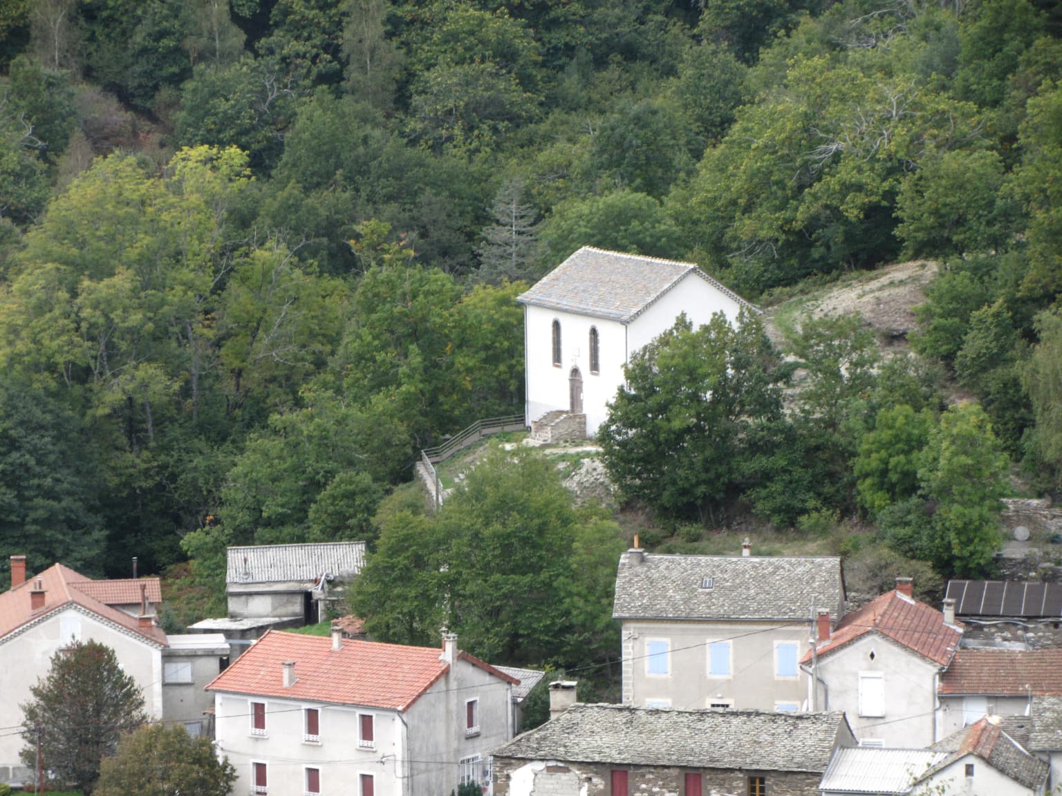 White building with gray roof on a hill surrounded by green trees and village buildings with red roofs
