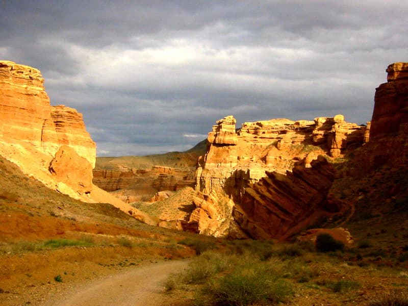 Red rock canyon with layered rock formations, winding dirt road, sparse vegetation, and overcast sky