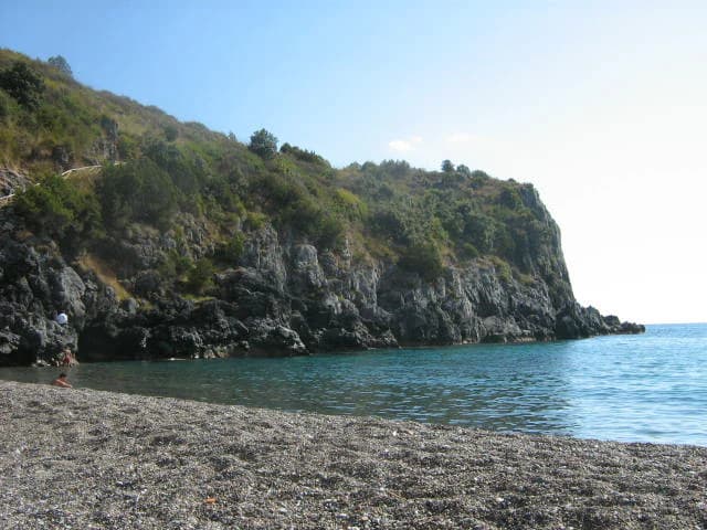 Pebbly beach with turquoise water, rocky cliffs covered in green vegetation, and a person walking along the shore under a clear blue sky