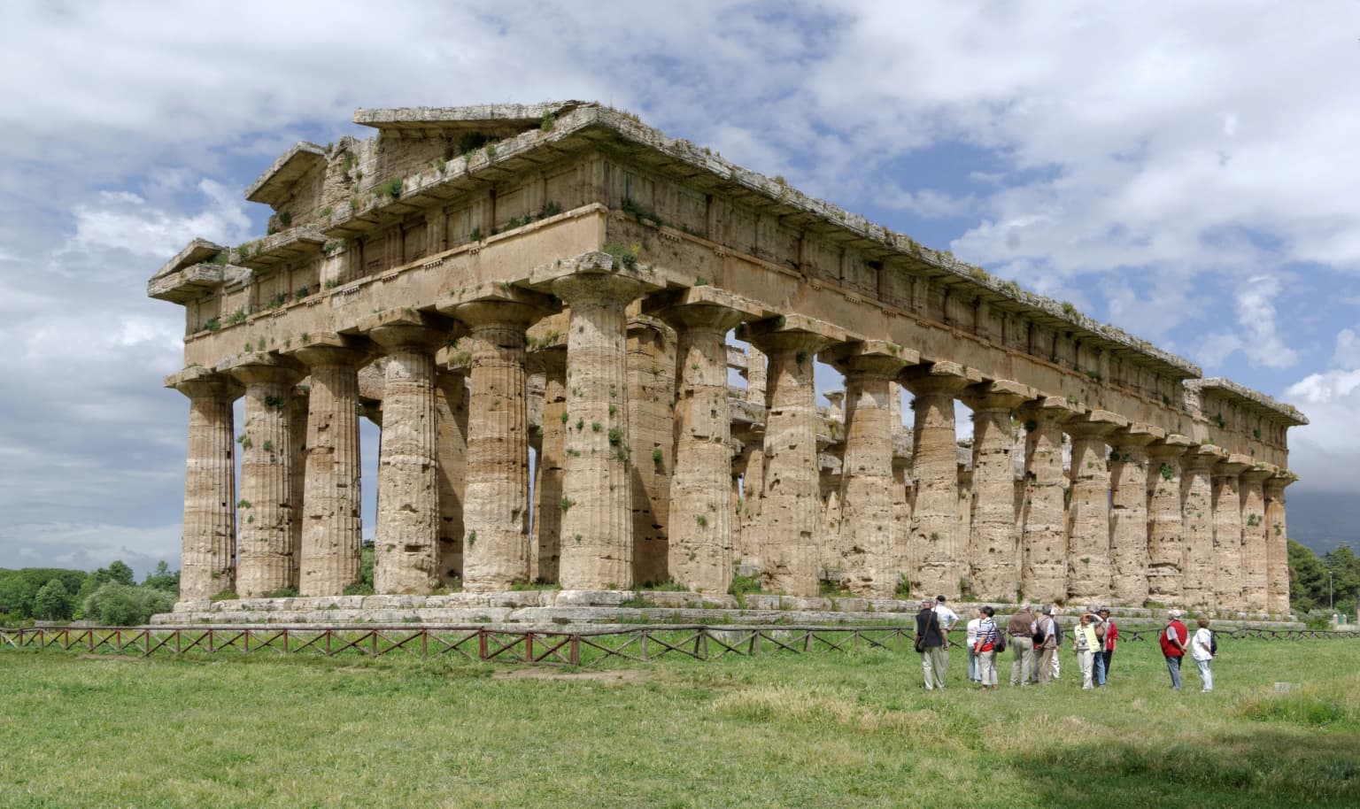 Ancient stone temple with tall columns, grassy field, and several people standing nearby under a partly cloudy sky