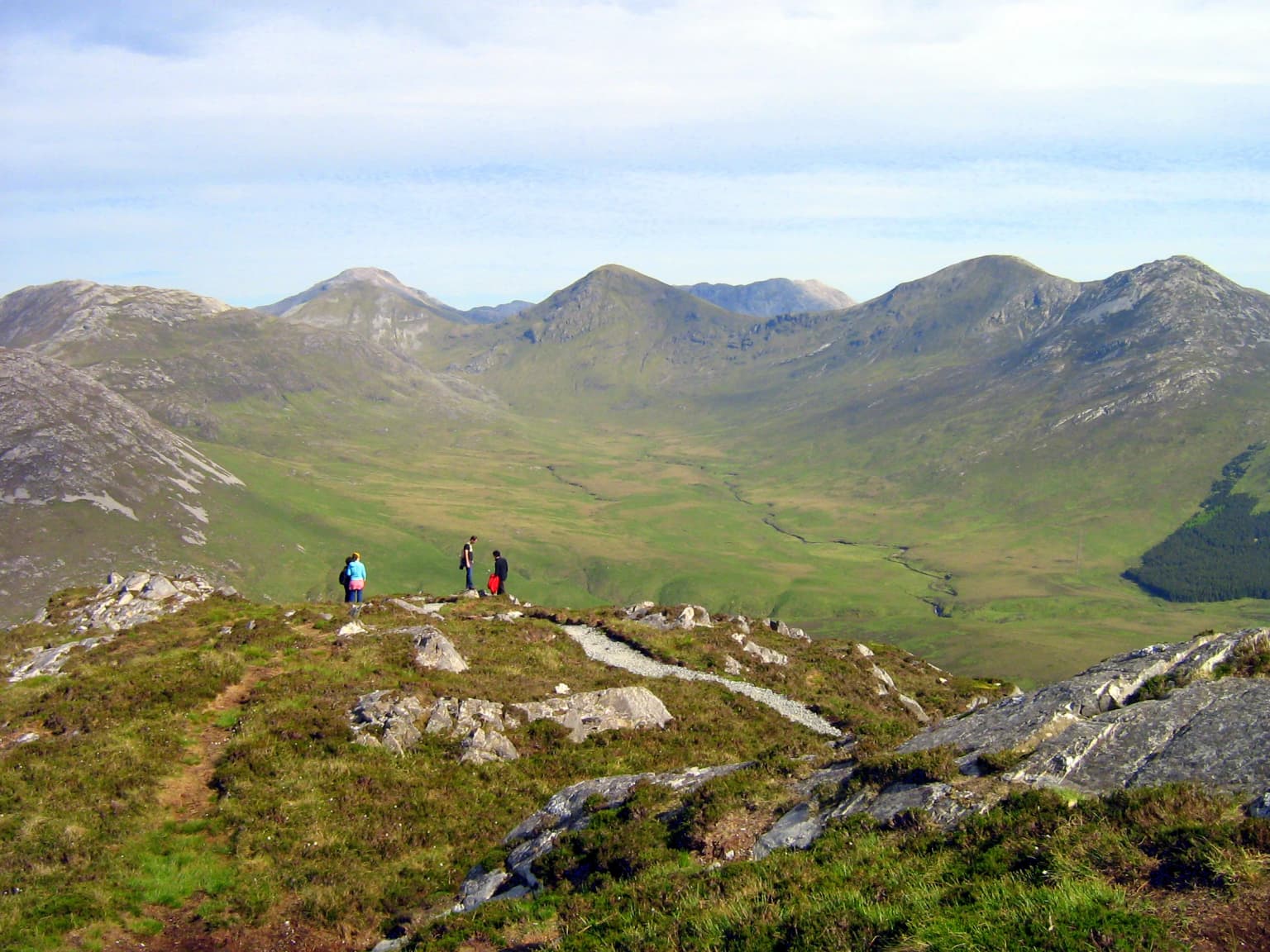 Three hikers standing on a rocky hilltop with green vegetation, overlooking a valley surrounded by mountain ranges under a partly cloudy sky
