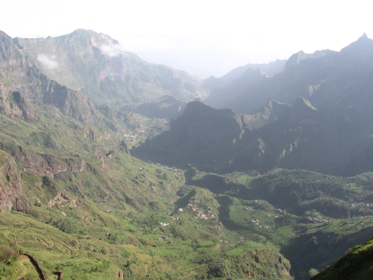 Vast green valley with rugged mountain slopes, scattered settlements, and winding paths viewed from a high vantage point