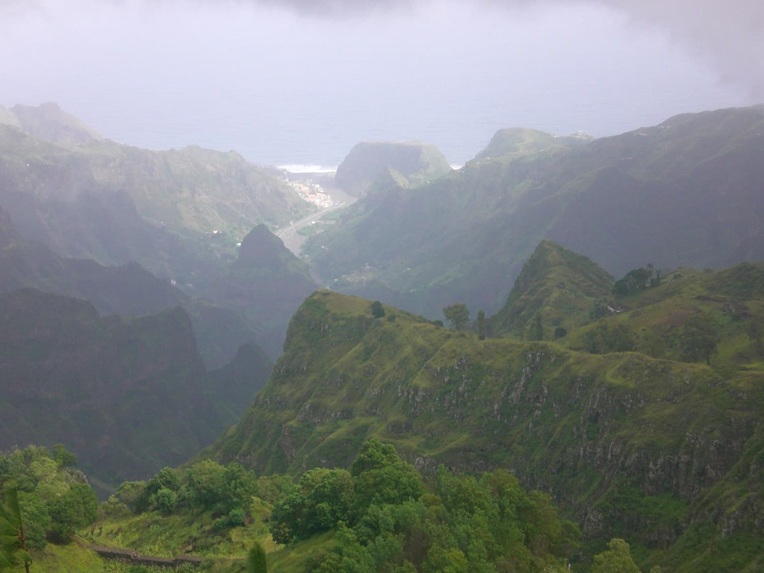 Wide landscape of lush green mountain slopes, valley, and distant city under overcast sky