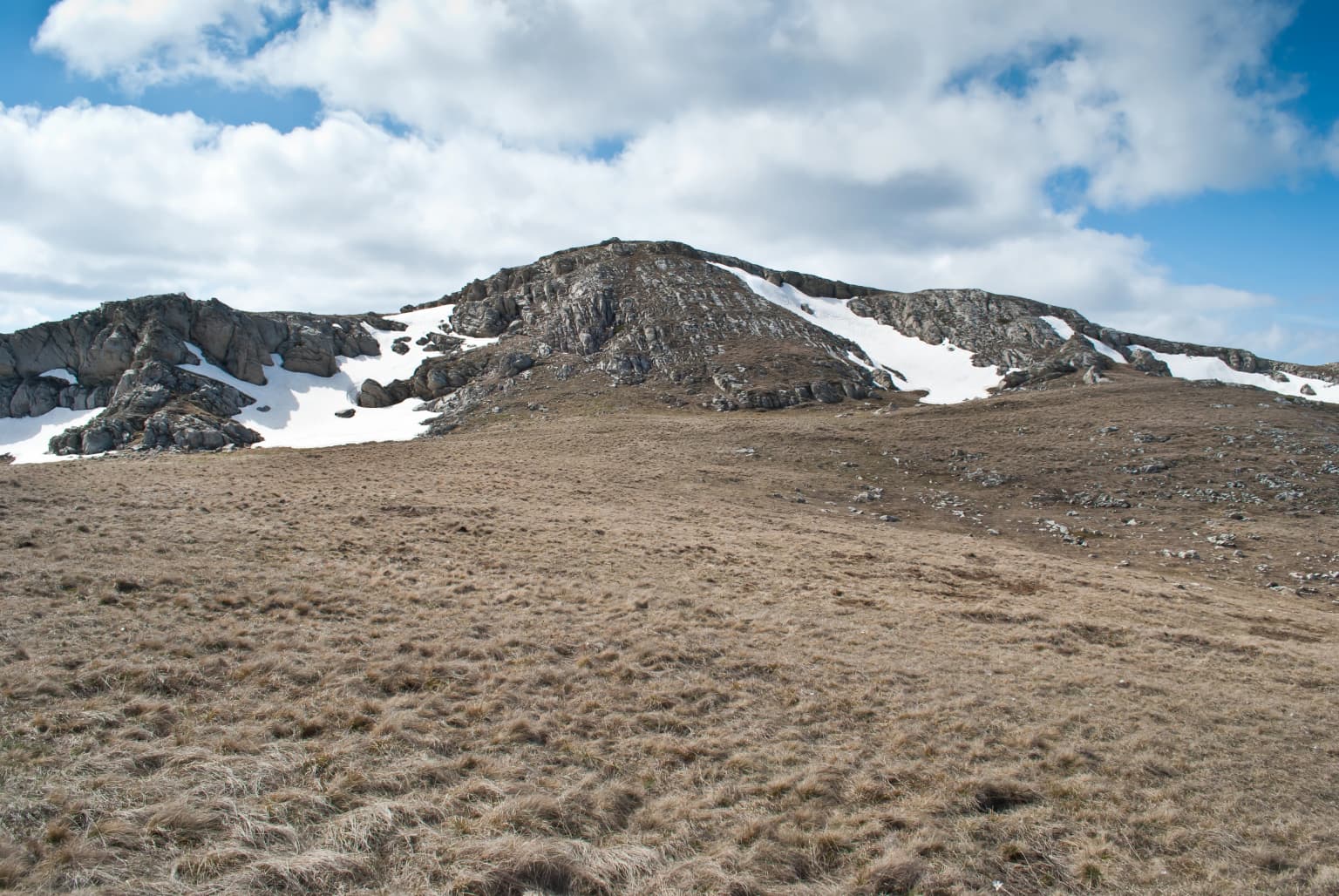 Grassy plain leading to rocky mountain slopes with snow patches under a partly cloudy sky