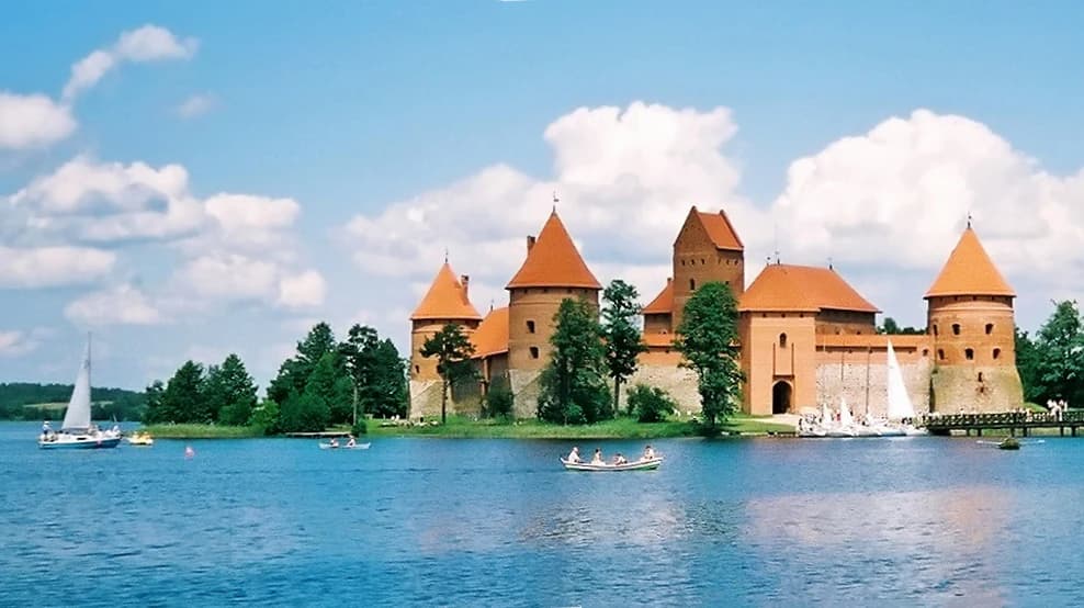 A wide shot of a red brick castle with orange conical roofs on an island in a blue lake, with sailboats and rowboats on the water under a blue sky with clouds.