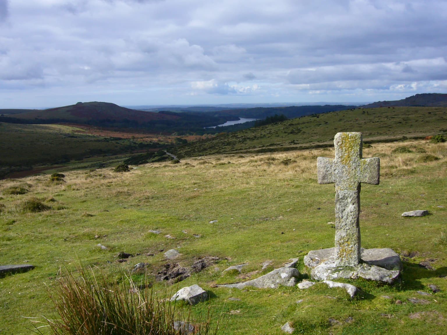 Stone cross standing on grassy moorland with distant hills and reservoir under cloudy sky