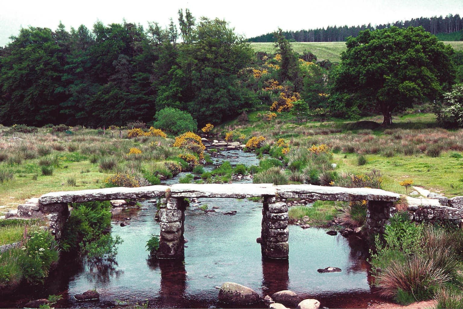 Stone clapper bridge spanning a river with green moorland and trees in the background
