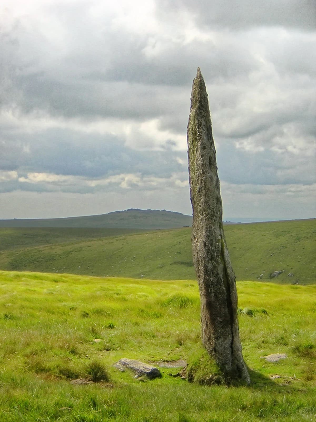 Tall weathered standing stone in a grassy moorland field with rolling hills under a cloudy sky