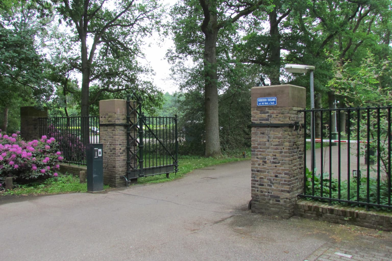 Park entrance gate with brick pillars and black iron fencing, paved road, greenery and trees in background