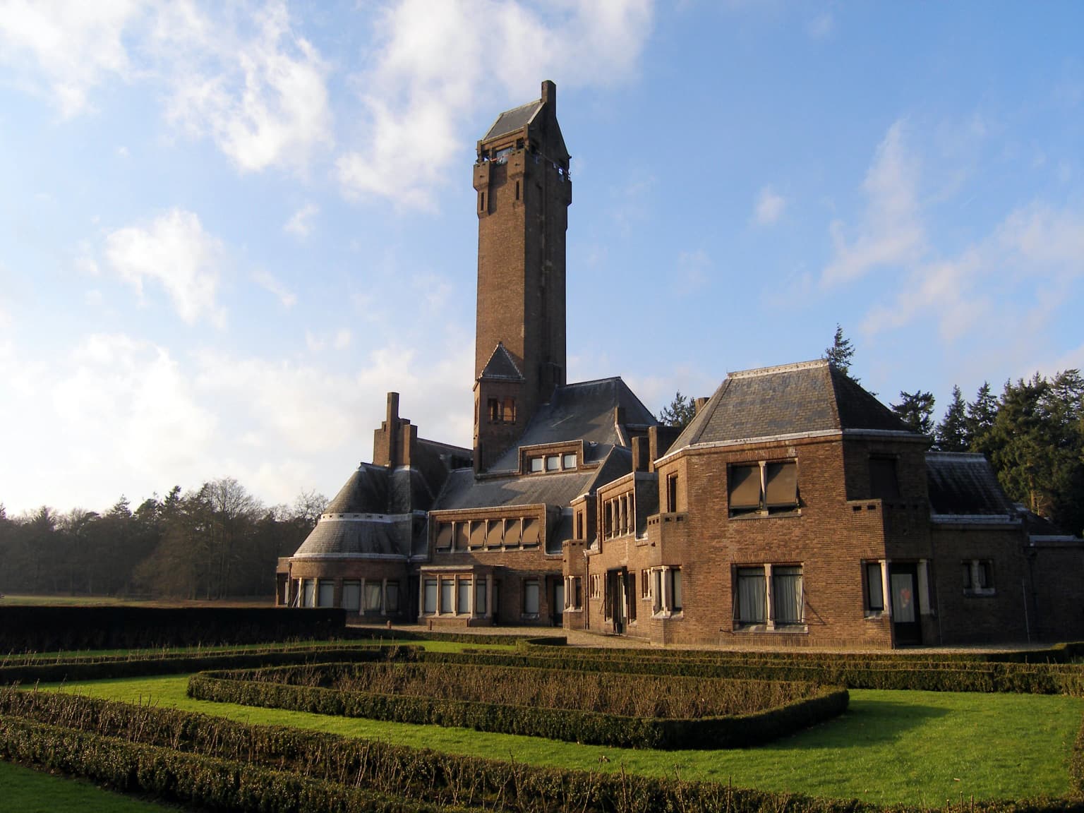 A large brick building with a tall tower and manicured gardens under a partly cloudy sky