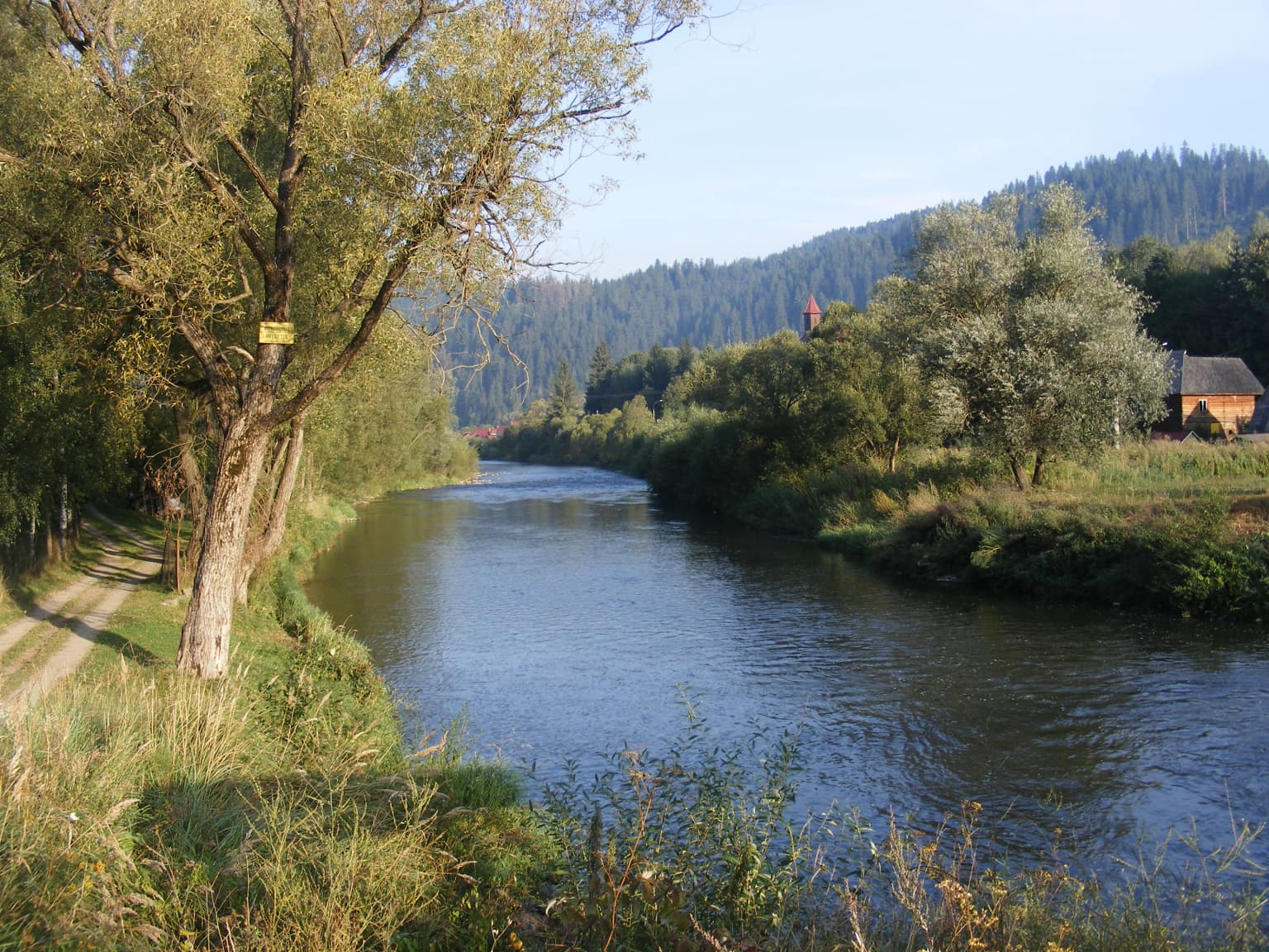 A river flows through a valley bordered by trees, with a dirt path on the left and a small building on the right under a clear sky.