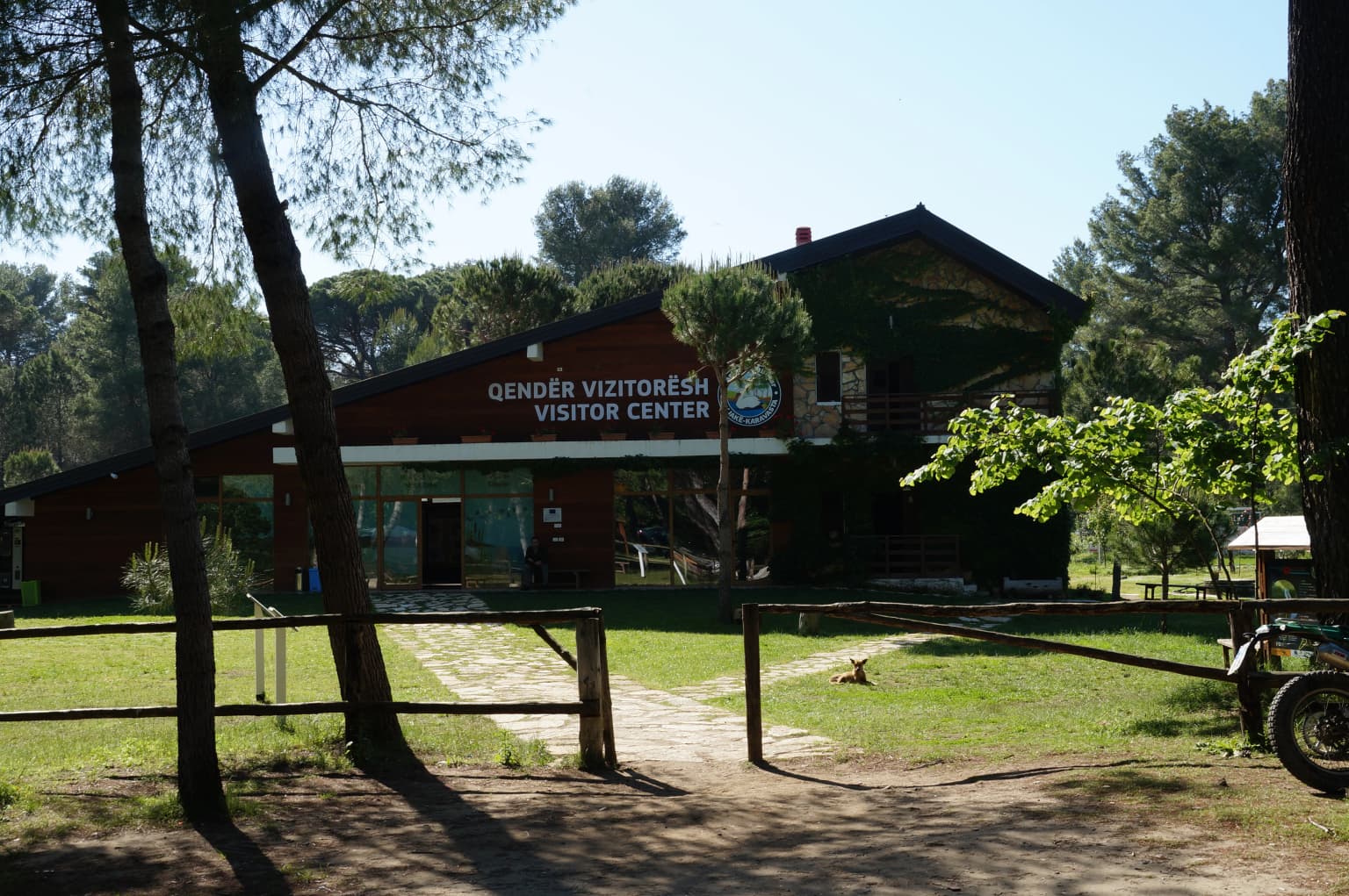 Wooden visitor center building with sign reading 'GJENDËR VIZITORESH VISITOR CENTER' surrounded by trees and a grassy area with a wooden fence