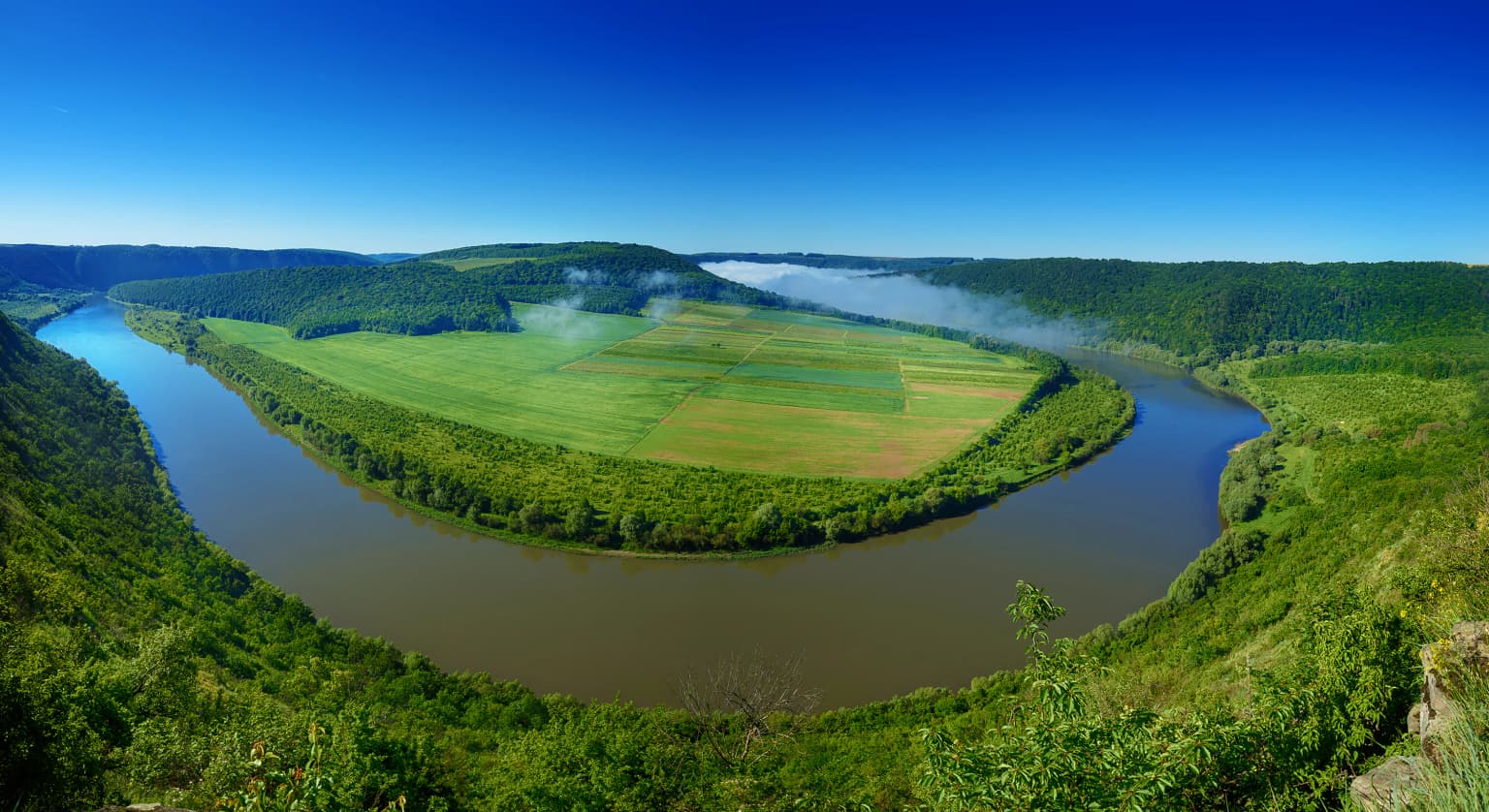 Wide panoramic view of a winding river surrounded by green fields and forests under a clear blue sky