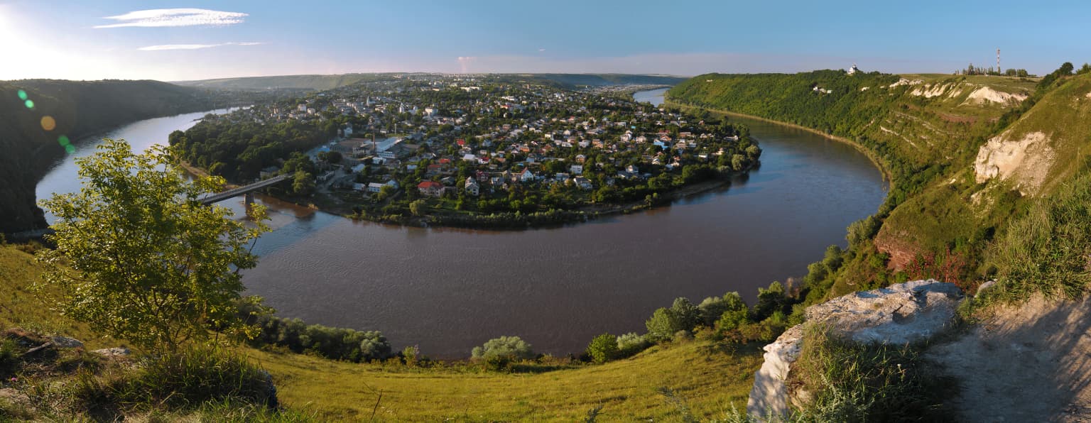Wide-angle view of the Dniester River curving through a canyon, with a small town nestled on the riverbank, surrounded by rolling hills and greenery