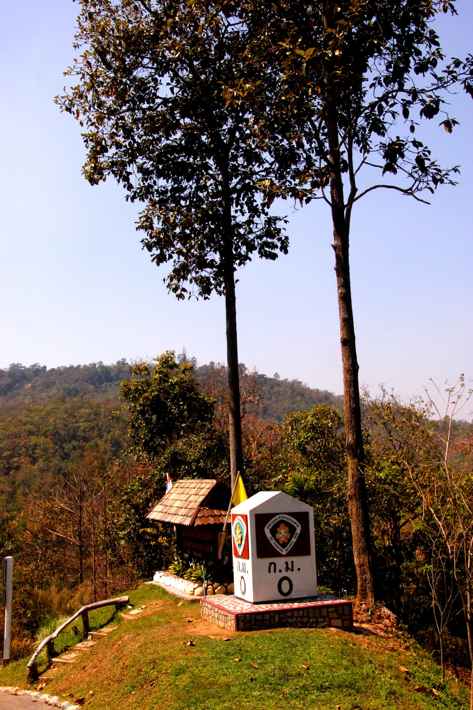 A concrete park entrance sign with Thai text and emblem surrounded by trees, a small hut, and forested hills in the background