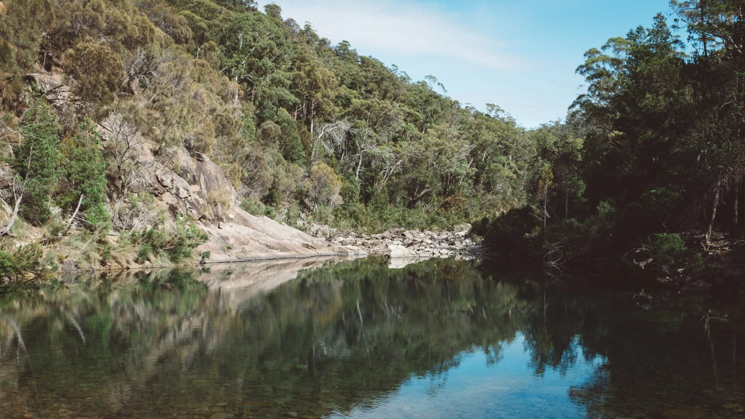 Calm river reflecting rocky hills and dense forest under clear sky