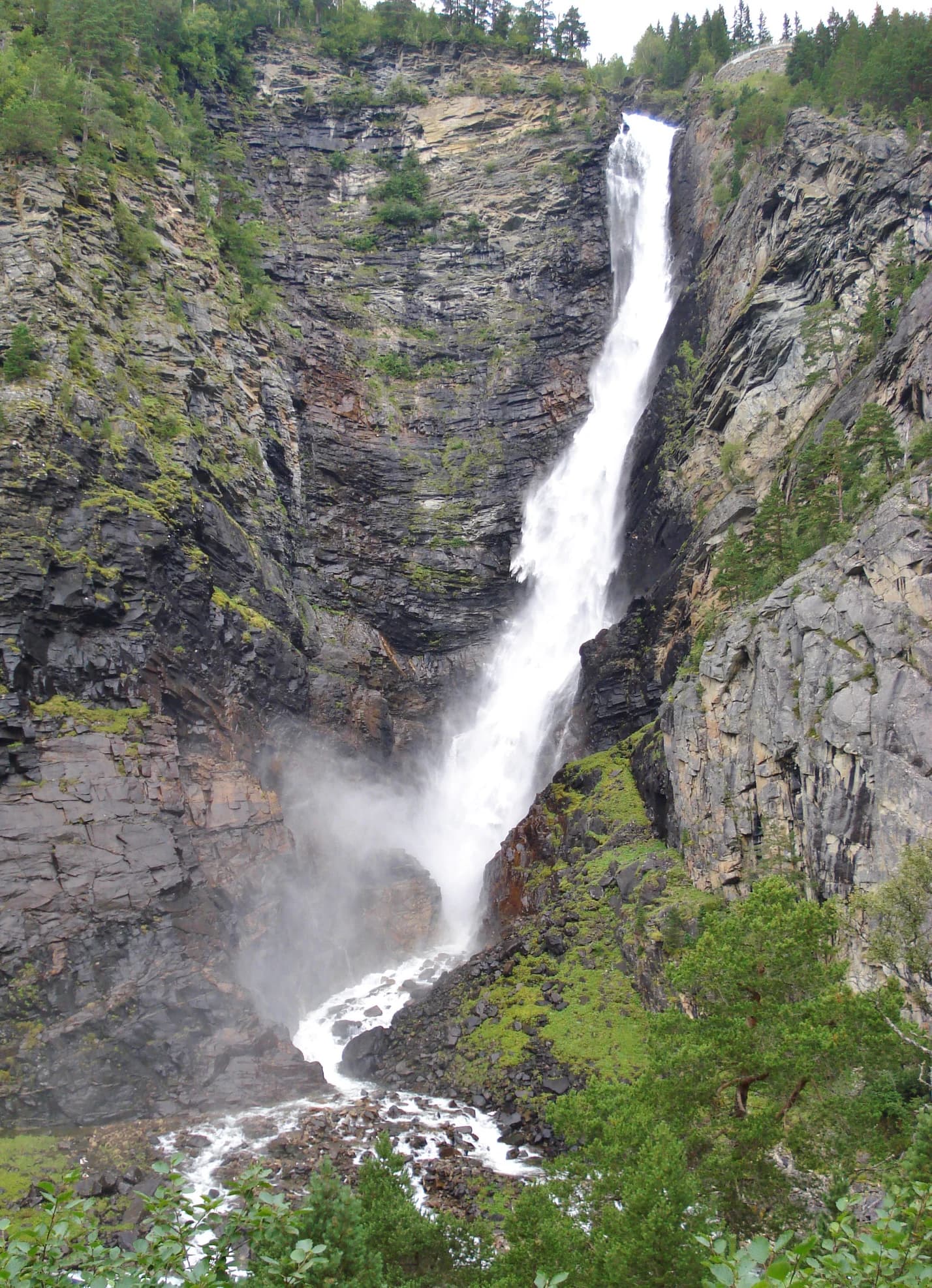 Tall waterfall flowing down a steep rocky cliff surrounded by green vegetation and trees.