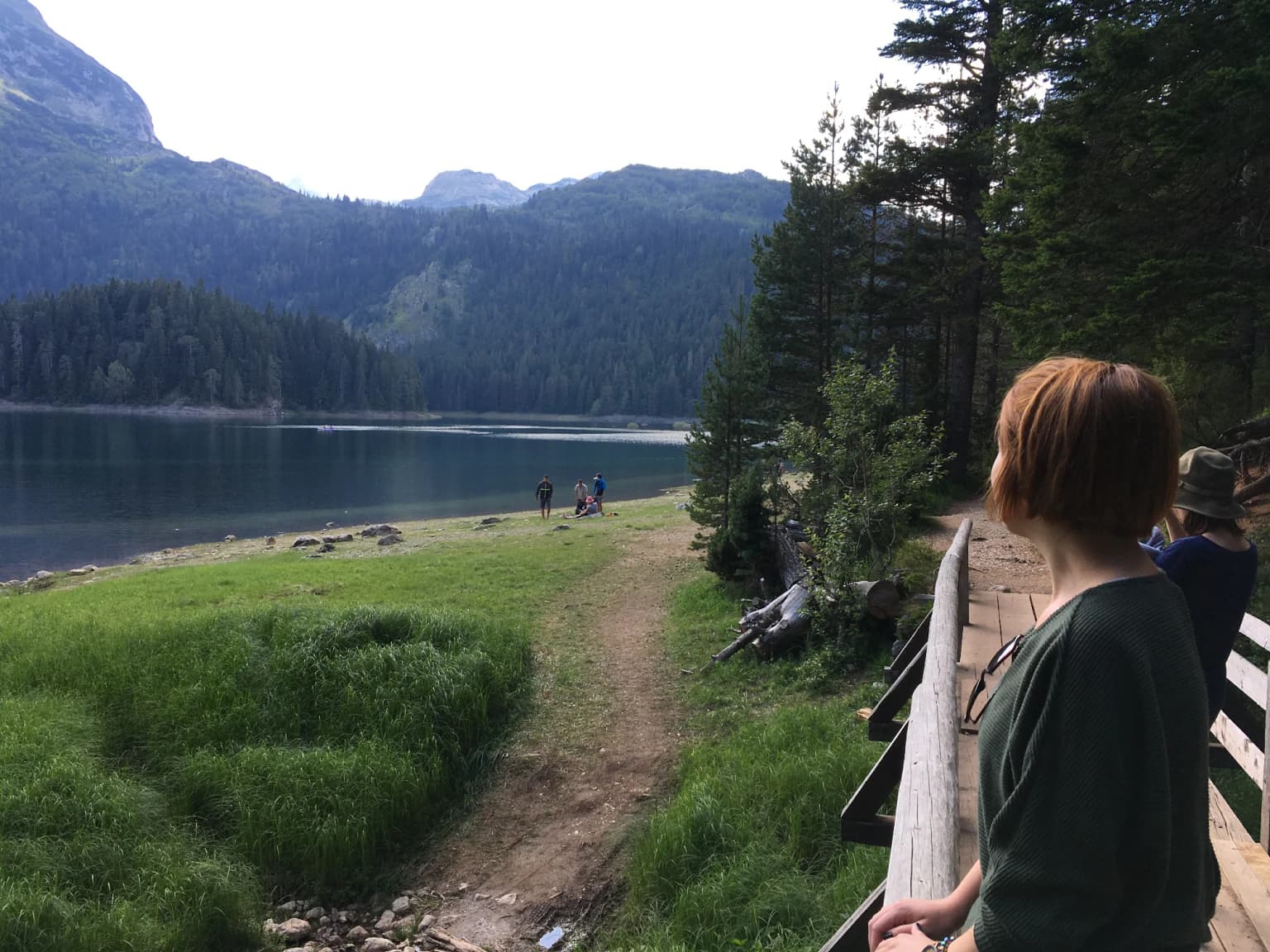 A lake surrounded by mountains and forest, with a wooden boardwalk and several people walking along a path