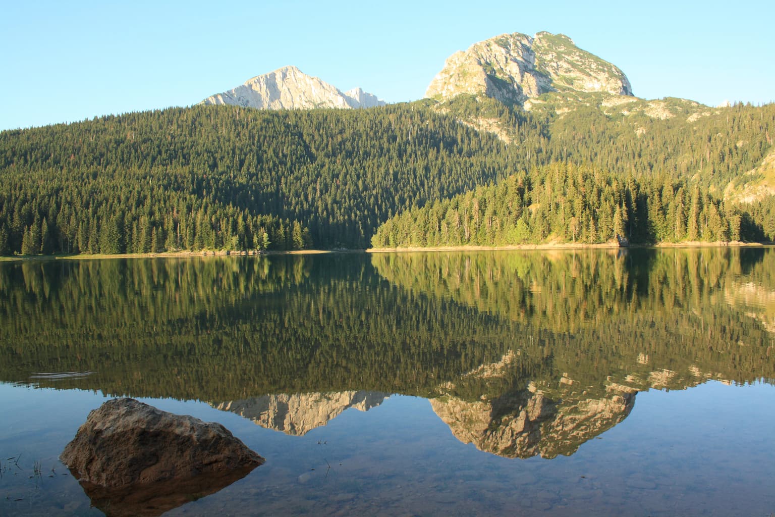 A calm lake reflecting mountains and forests with a large rock in the foreground