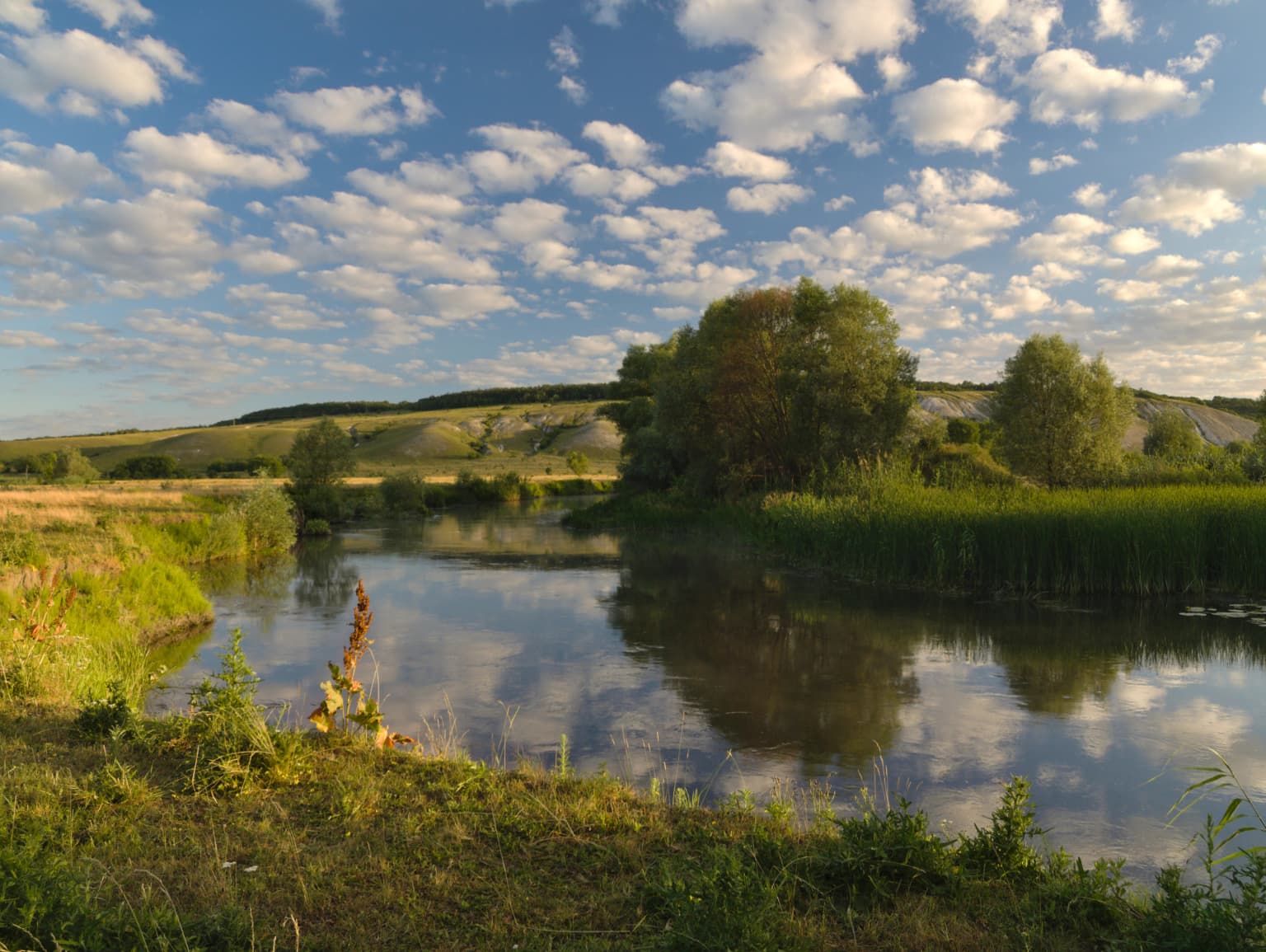 Calm river reflecting blue sky with scattered clouds, surrounded by green vegetation and rolling hills