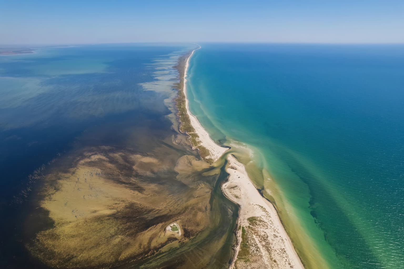 Aerial view of a narrow island with sandy beaches and clear blue water, showing the contrast between deeper ocean water and shallow coastal waters