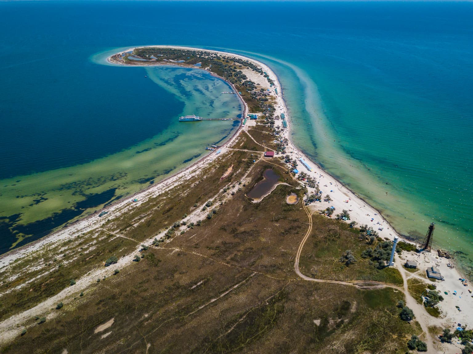 Aerial view of a narrow coastal peninsula with sandy areas, patches of vegetation, and shallow turquoise waters extending into the sea