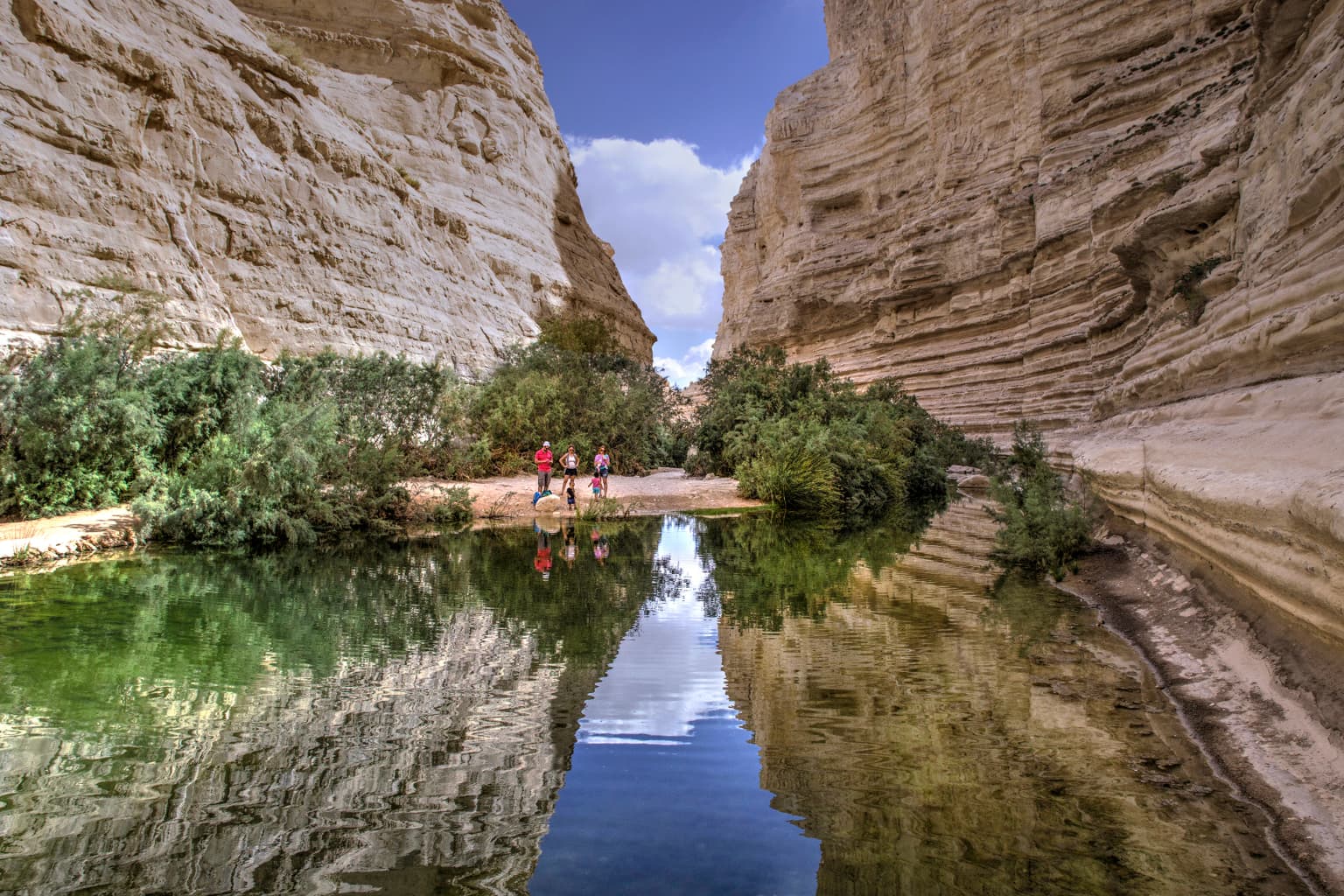Calm spring water reflecting rocky canyon walls, vegetation, and two people standing at the water's edge under a clear sky.