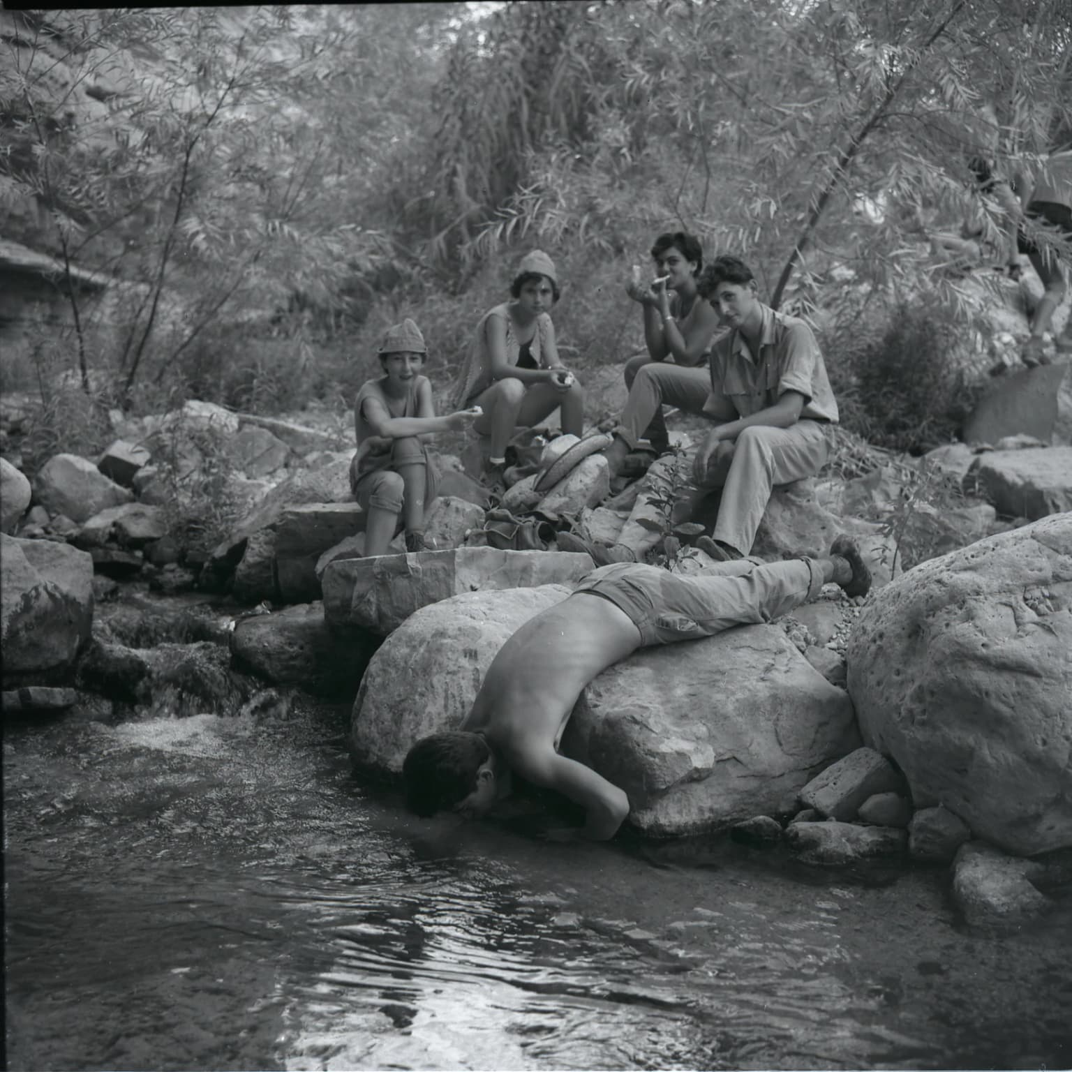 Black-and-white photograph showing several people near a stream in a rocky, vegetated area with vegetation in the background