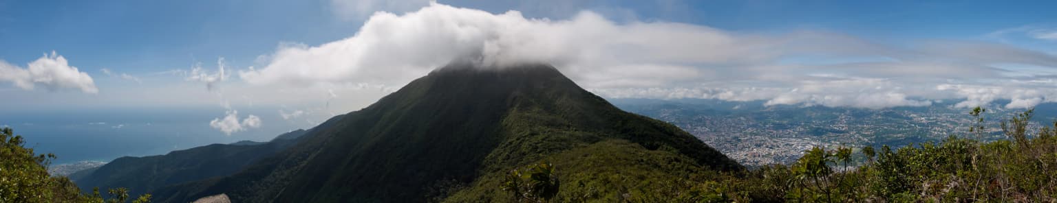 Panoramic landscape view of a mountain with vegetation, city in valley, and ocean under partly cloudy sky