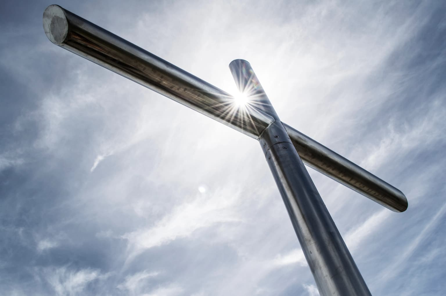 Metal cross structure with sun flare against a cloudy sky