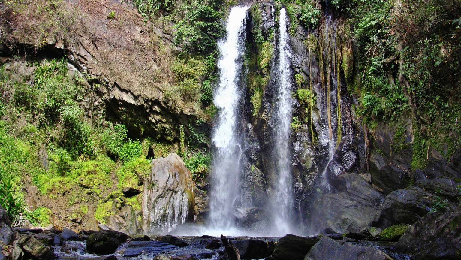 Cascada doble y alta que fluye sobre acantilados rocosos con densa vegetación verde a ambos lados