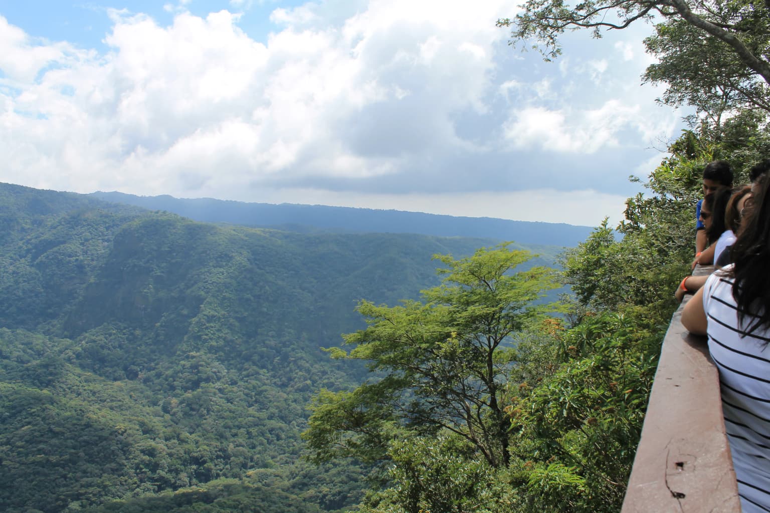 Dos personas de pie en una barandilla de madera con vistas a un paisaje montañoso boscoso con colinas verdes y nubes dispersas