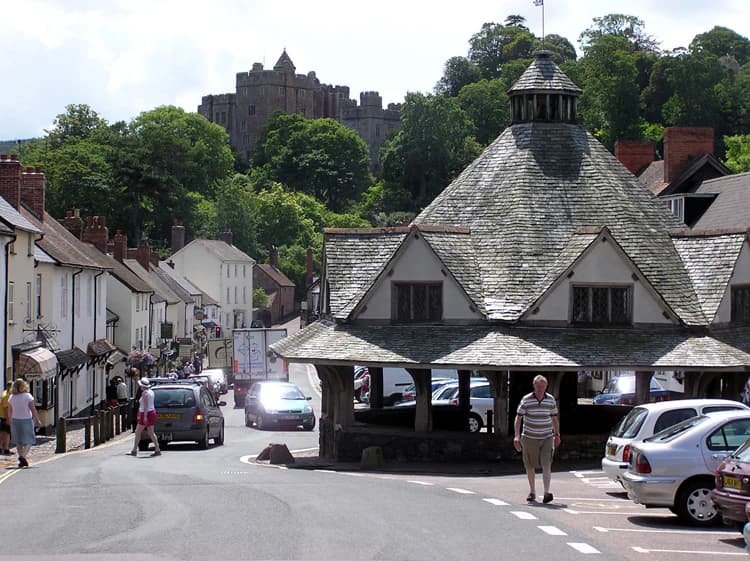 A street view showing the octagonal Yarn Market building with Dunster Castle visible on a hill in the background, surrounded by town buildings and pedestrians