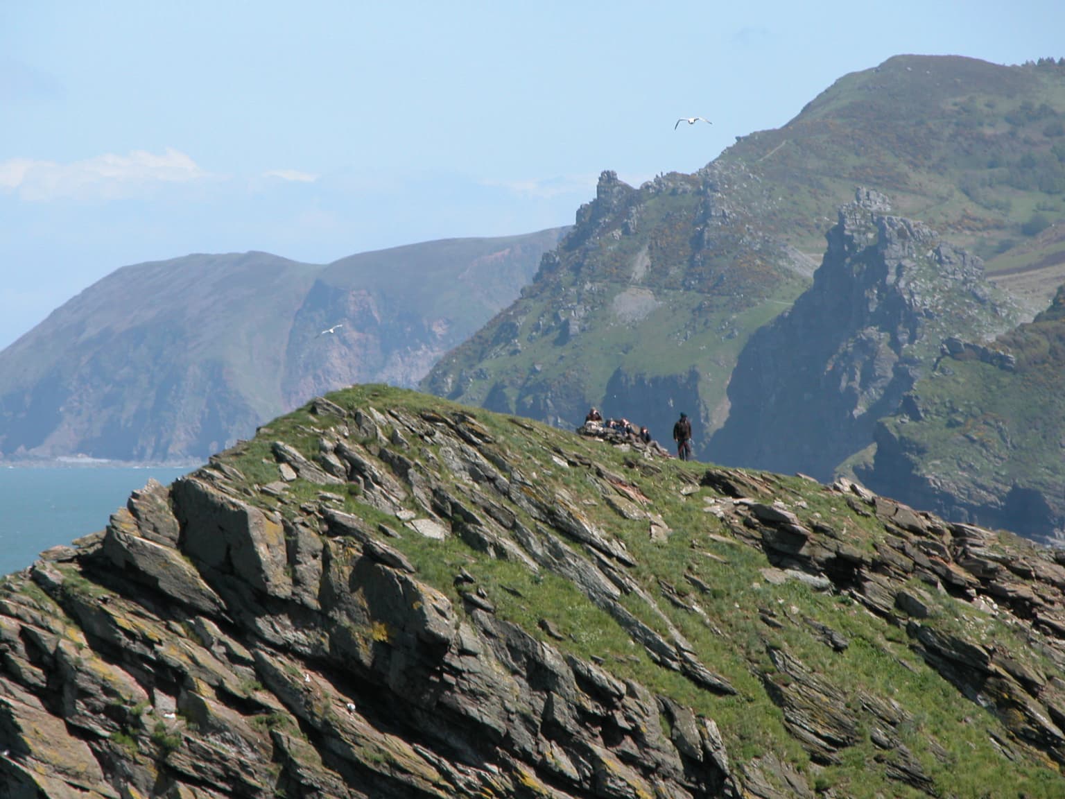 Rocky cliff with grassy patches and a small group of people standing at the summit, distant sea and hills under clear sky