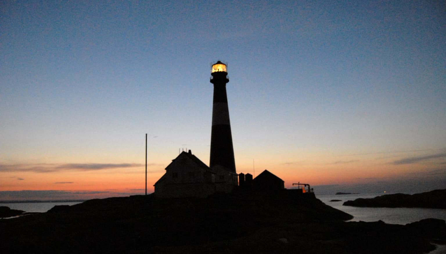 Silhouette of Færder Lighthouse with illuminated beacon at dusk, adjacent structures, and coastal waters under a gradient sky