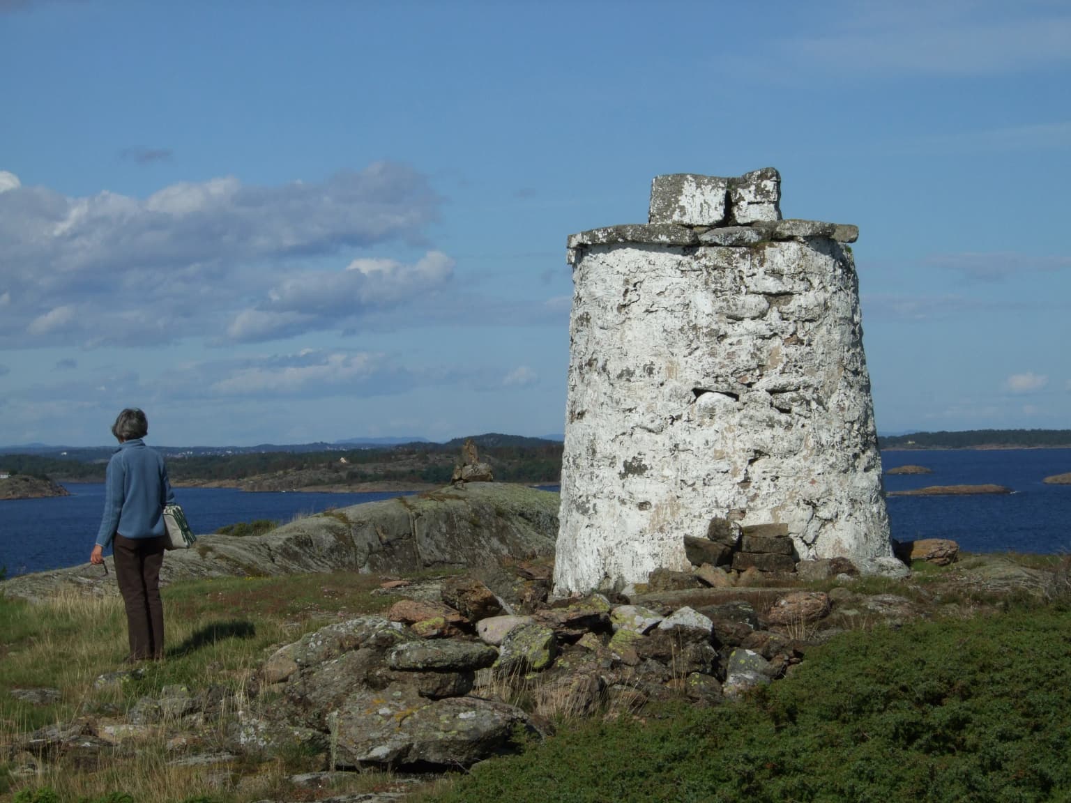 A person in a blue jacket standing on rocky terrain next to a tall white stone tower with water and islands in the background