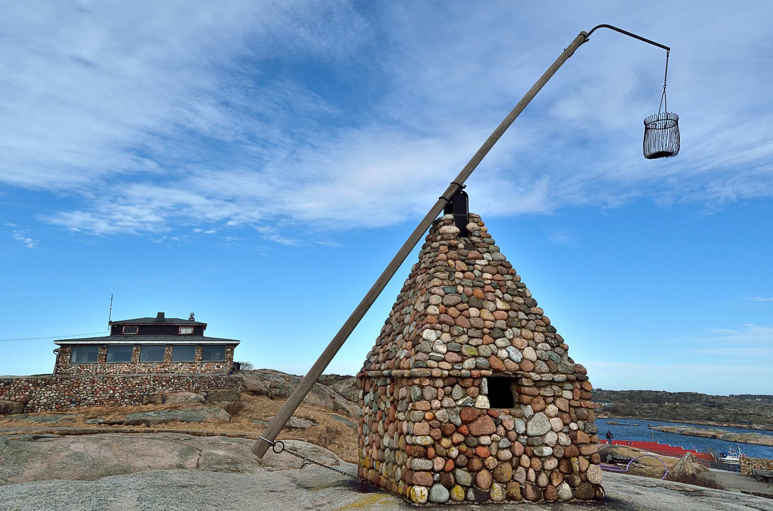 Stone tower with a wooden pole holding a metal basket, adjacent to a multi-story building by the water, under a blue sky with scattered clouds