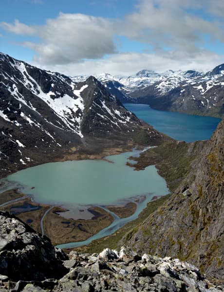 Panoramic view of a turquoise mountain lake surrounded by rocky terrain and snow-capped peaks under a partly cloudy sky