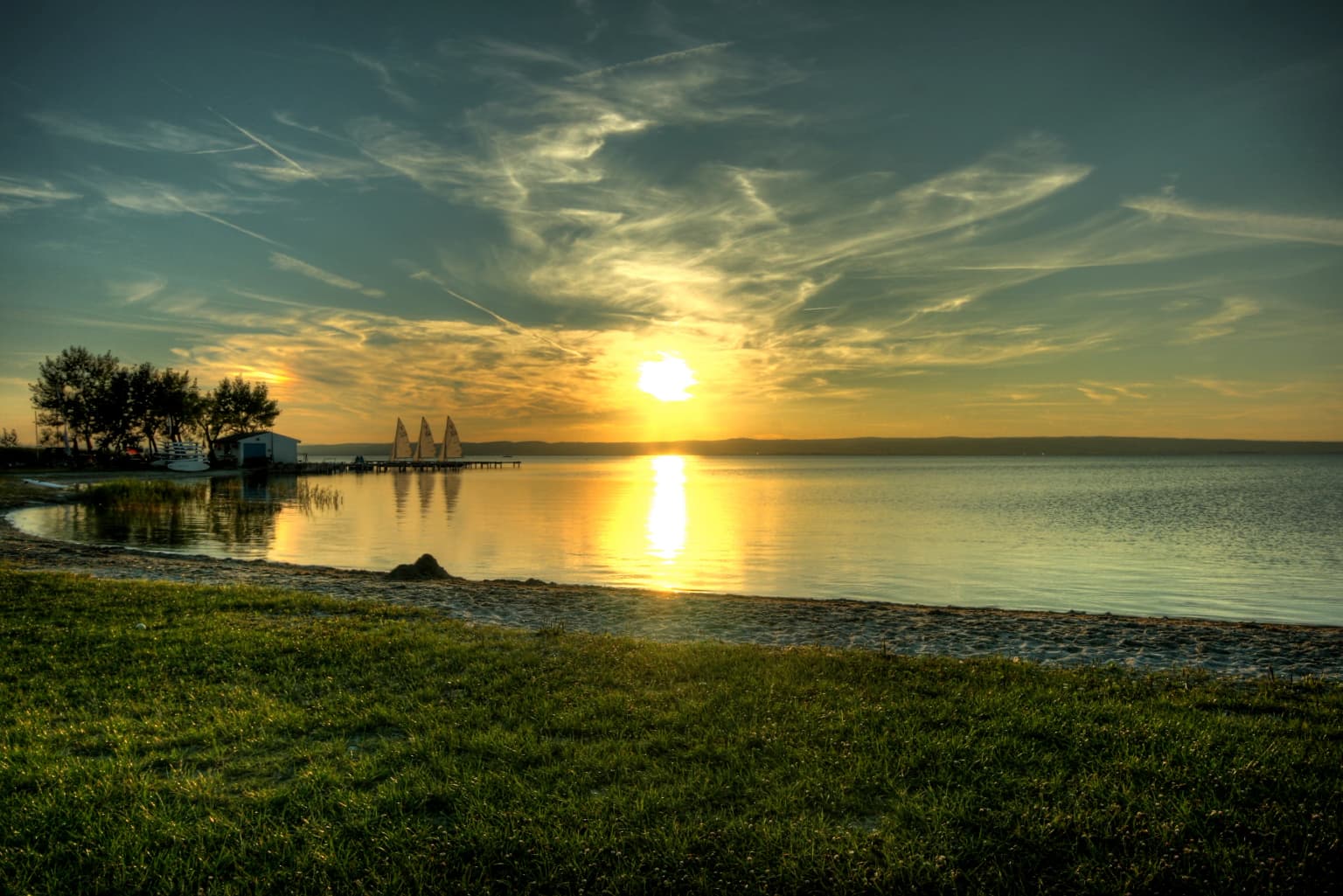 Sunset over a large lake with golden reflection on water, grassy shoreline in foreground, trees and small structure on left, clear sky with scattered clouds