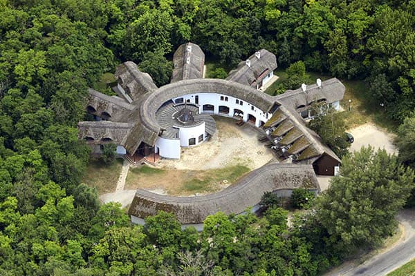 Aerial view of a circular building with multiple thatched roofs surrounded by dense green forest