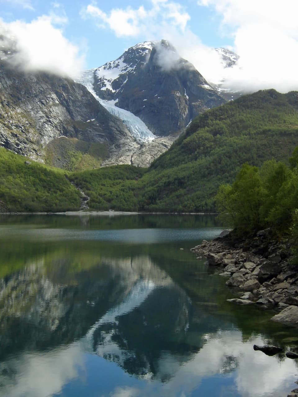 A lake reflecting a mountain range with a glacier front, surrounded by green valleys and rocky shoreline