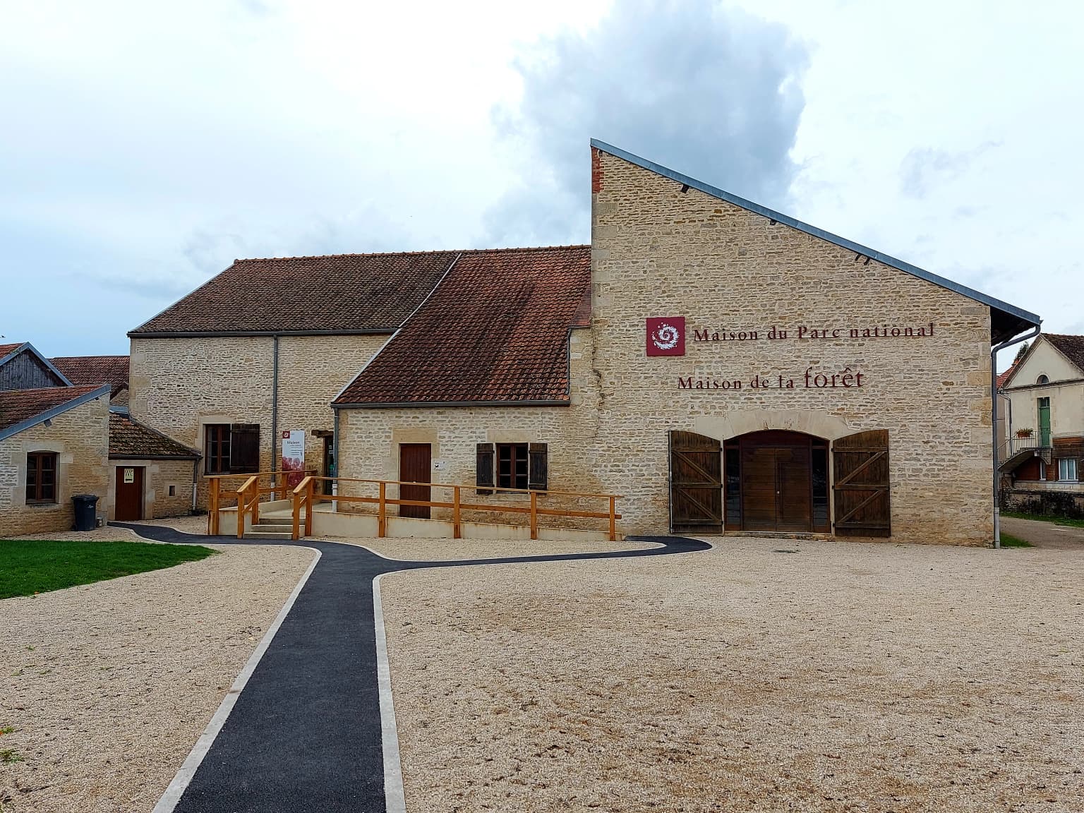Stone building with brown roof and red circular emblem, labeled 'Maison du Parc national de forêts' and 'Maison de la Forêt,' with gravel courtyard and paved pathway