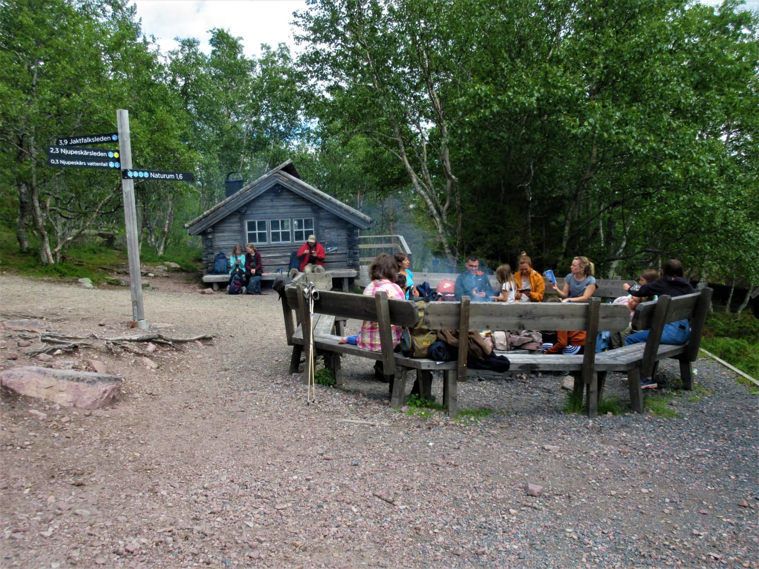 Group of people seated at a circular wooden bench in a gravel rest area with a signpost, small cabin, and forested landscape in the background