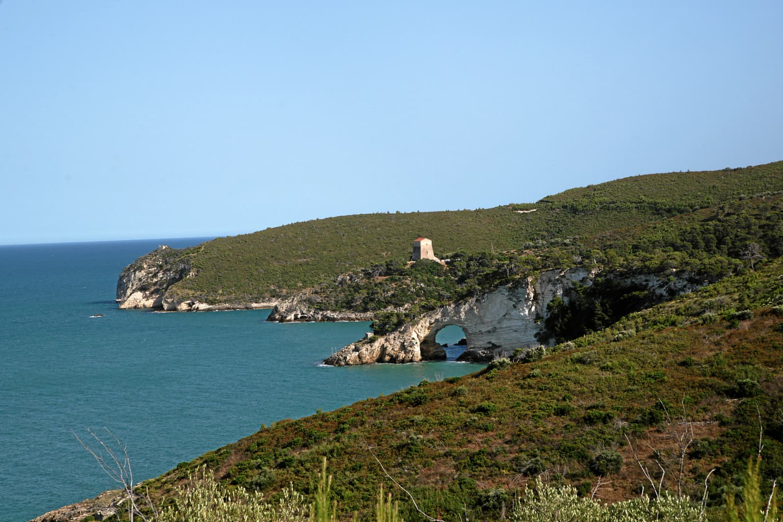 Coastal landscape featuring turquoise sea, rocky cliffs, a natural arch, and a small tower on a hillside