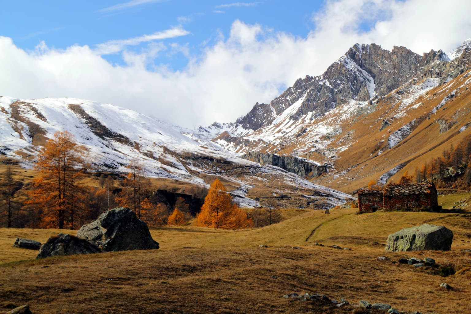 Autumn landscape featuring snow-covered mountain peaks, golden larch trees, rocky terrain, and a small stone structure under a partly cloudy sky