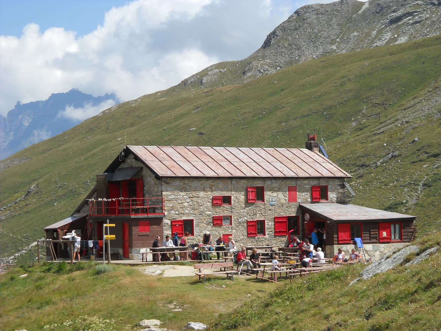 Stone building with red windows and metal roof, surrounded by grassy mountains, with people gathered on a terrace.
