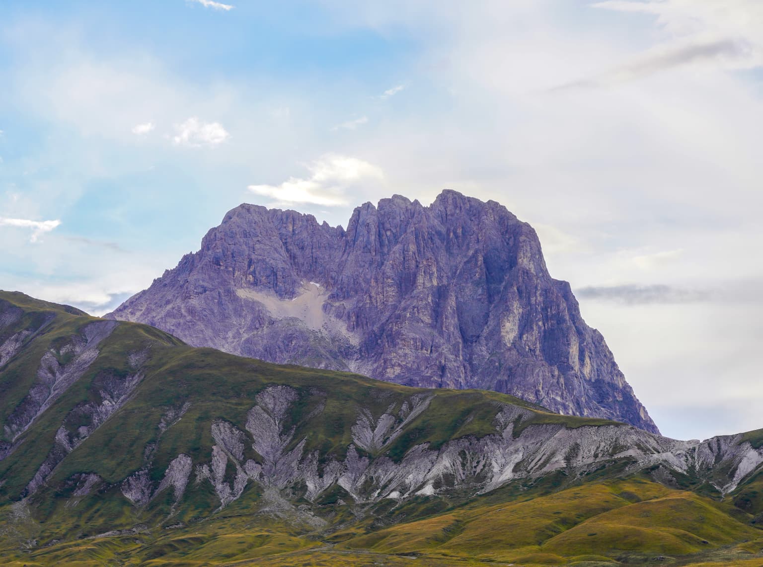 Rugged mountain peak with rocky cliffs and green slopes under a partly cloudy sky