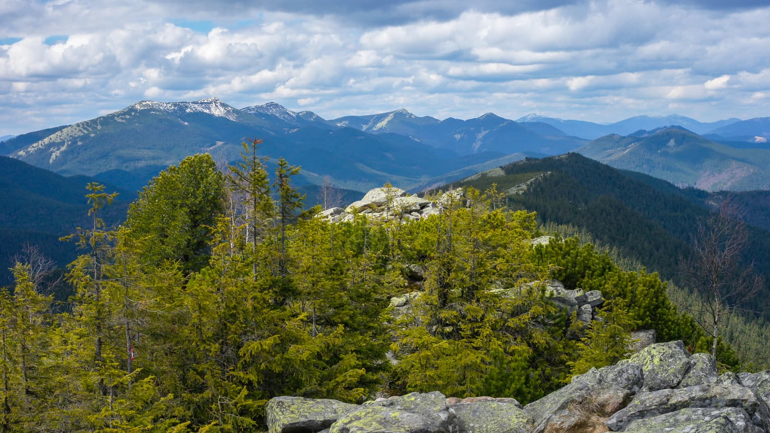 Panoramic view of mountain peaks with patches of snow, forested slopes, and rocky terrain under a partly cloudy sky
